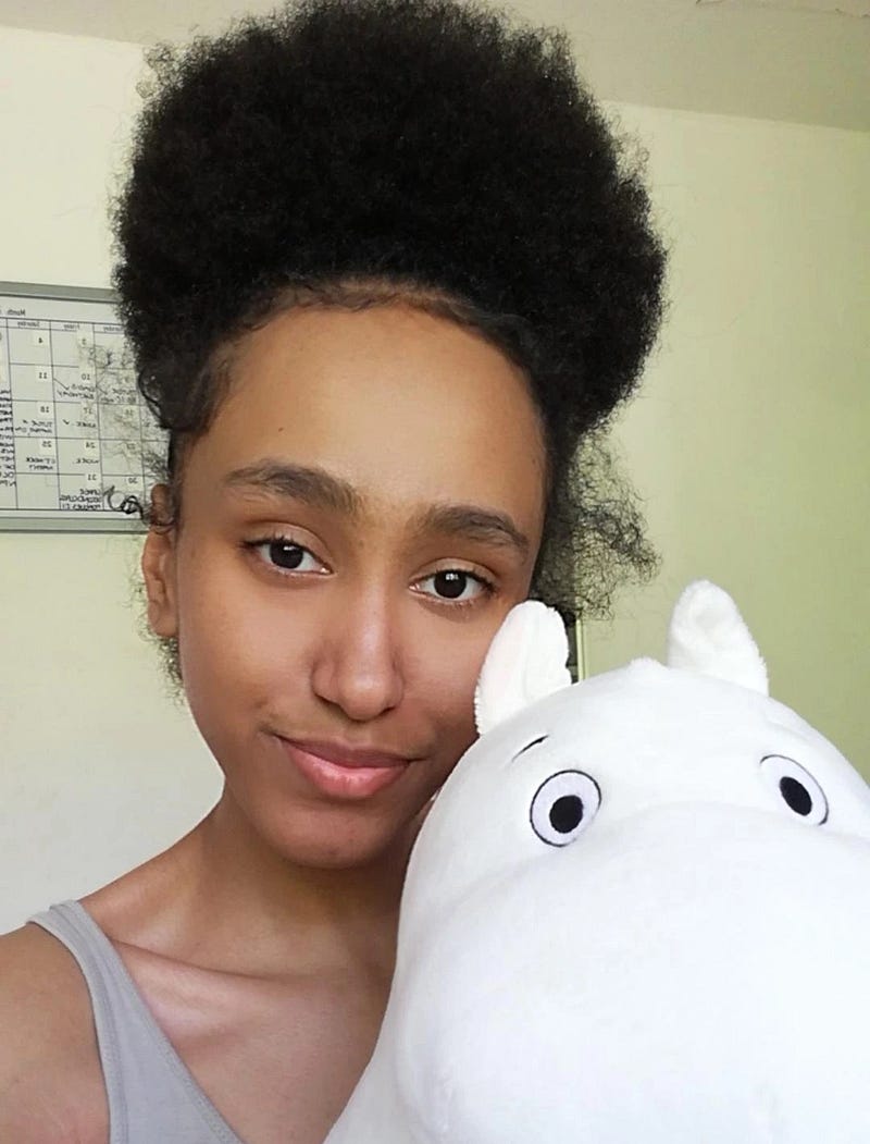 A recent photograph of me (Kashala, aged 21) proudly wearing my natural hair, posed with a Moomin plushie.