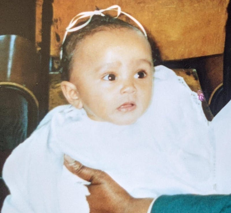 A photograph of me (Kashala, aged around 6 months or so) as a baby, dressed in white, waiting to get christened.
