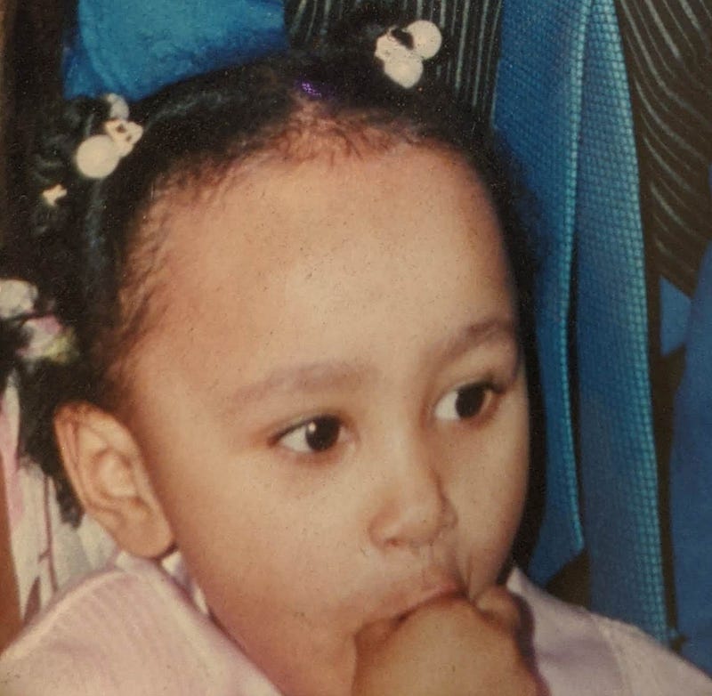 A photograph of me (Kashala, aged around 3 or so) as a child, dressed in pink, with my fingers in my mouth.